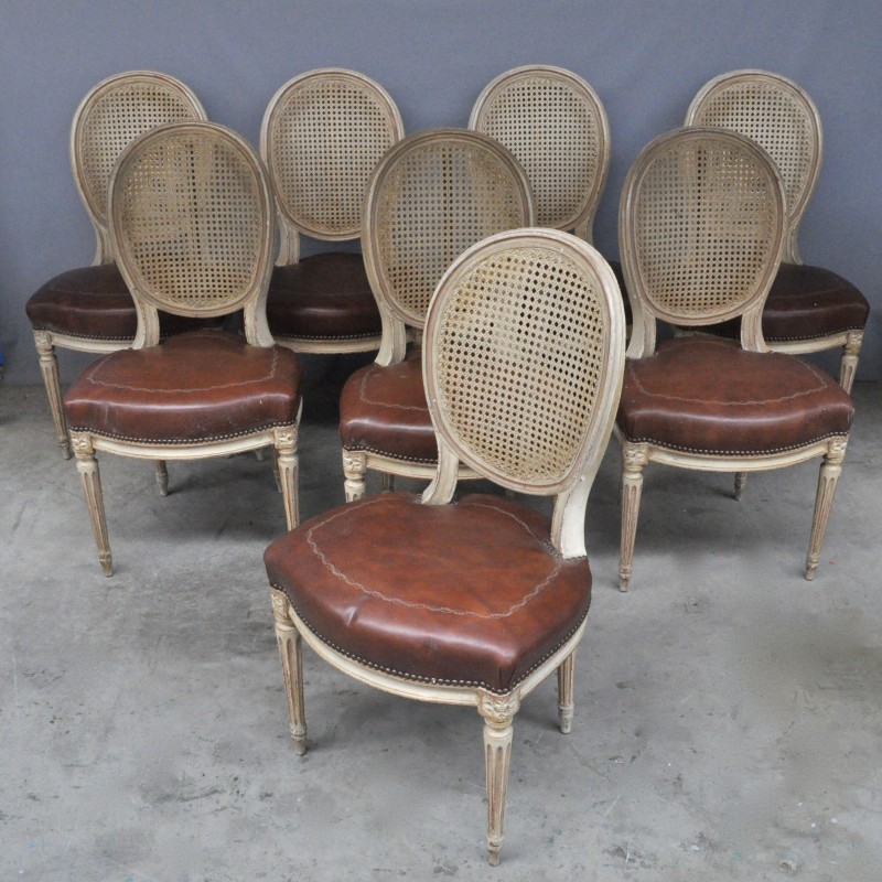 Een serie van acht witgelakte stoelen in Lodewijk XVI-stijl. Ruggen gevlochten, zitten bekleed met bruin leder. Gecanneleerde poten.