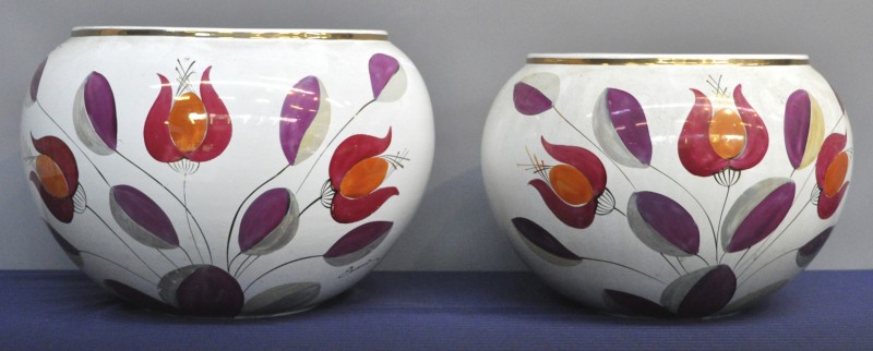 Een paar bolle cachepots (de éne iets groter dan de andere) met een veelkleurig gestileerd bloemenmotief. Met de hand geschilderd en gesigneerd Emanuela. Jaren zestig.