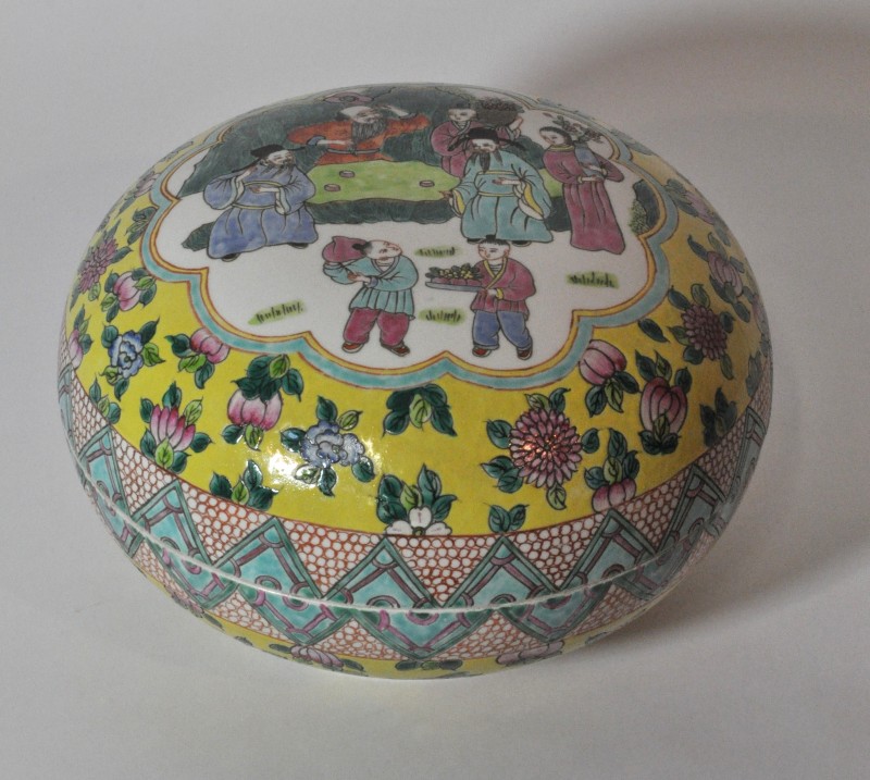 Een ronde, platte doos van polychroom porselein met decor van personages en bloemen. Chinees werk.