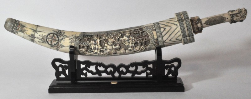 Een mes in schede van gesculpteerd en deels gepolychromeerd been. Chinees werk op houten staander.