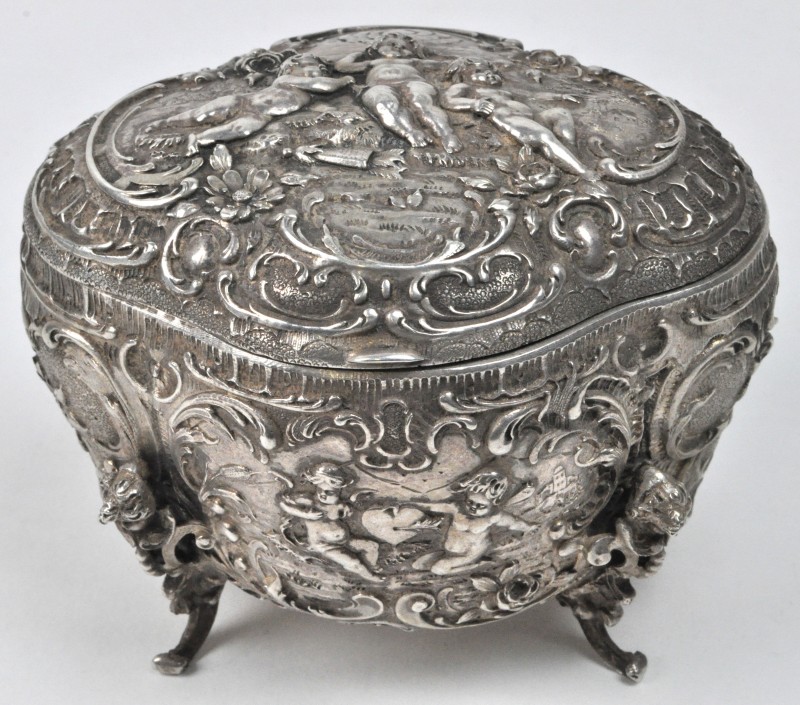 Een dekseldoosje van gedreven zilver in barokke stijl op vier pootjes, versierd met putti in reliëf. Binnenin met vermeil.