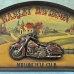 “Harley Davidson Motorcycle Club”. Een houten reclamepaneel met een motorfiets in reliëf. Naar de jaren ‘50.