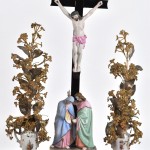 Een kruisbeeld met Maria en Johannes van meerkleurig biscuit op een porseleinen sokkel en met bijhorende vaasjes. Onder stolp.