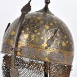 Een Perzische Kulah Khud helm met een gedamasceerd druivendecor en koranverzen op zwarte fond versierd. Boven op de bolle helm een vierhoekige punt en twee aigrettehouders en achteraan een maliën lap. Omstreeks 1900.
