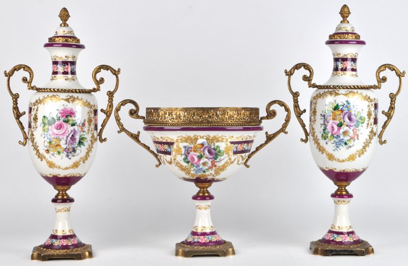 Een driedelig schouwstel van porselein en messing, bestaande uit een coupe en twee siervazen met een handgeschilderd bloemendecor. Naar de XVIIIe eeuw. Schade aan de voet van één van de vazen.