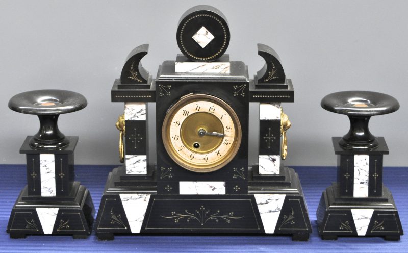 Een driedelig klokstel, bestaande uit een pendule en twee cassolettes van zwart en grijsgeaderd marmer en aan weerskanten versierd met een messingen leeuwenkop. Omstreeks 1900. Glas voor wijzerplaat manco.