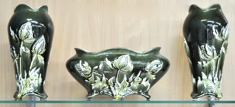 Een driedelig kaststel, bestaande uit een jardinière en twee vazen van meerkleurig aardewerk, versierd met een disteldecor in reliëf. Onderaan gemerkt.