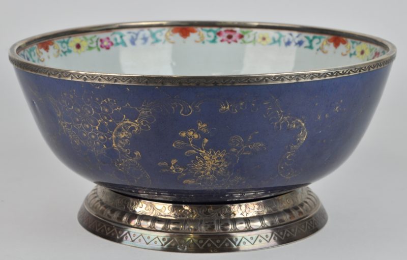 Een XIXe eeuwse kom van Chinees porselein met een (vervaagd) verguld decor op een blauwe fond, binnen versierd met een meerkleurig bloemenmotief op de rand en afgewerkt met 800‰ zilveren monturen met Brusselse keuren.