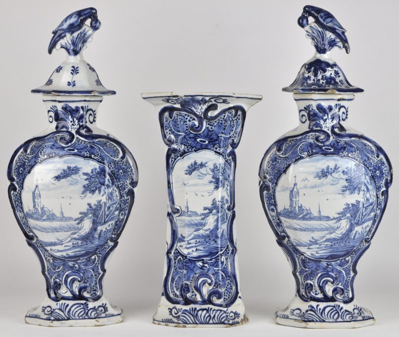 Een driedelig kaststel van blauw en wit aardewerk, gedecoreerd met een personage in een landschap. Restauraties aan één vaas, de dekselvazen in relatief goede staat. Onderaan gemerkt met de drie klokken. XVIIIe eeuw.