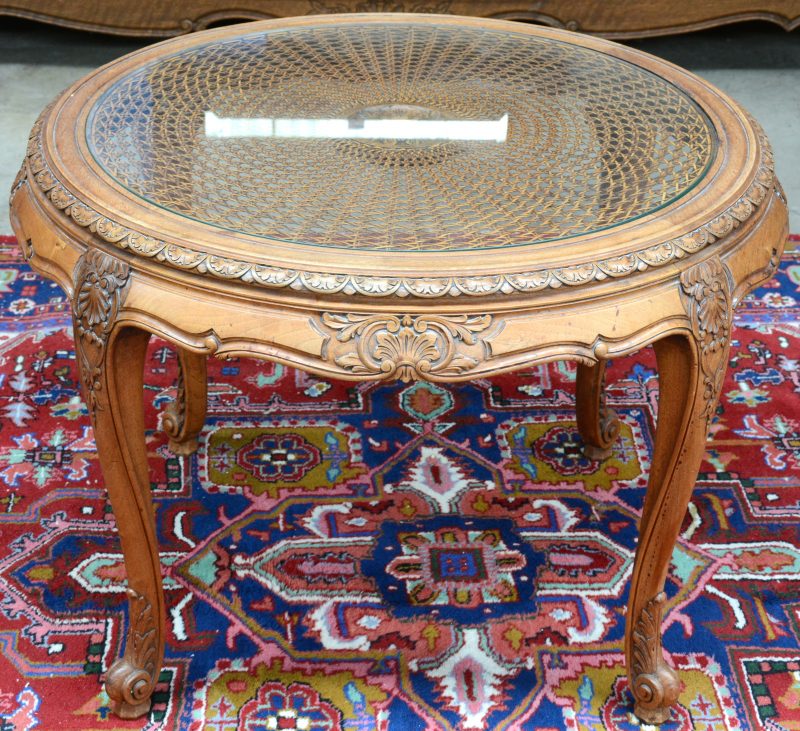 Een ronde salontafel van gesculpteerd notenhout met een gecanneerd blad onder glas.