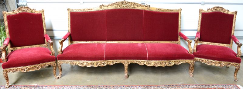 Een driedelig salongarnituur van verguld hout in Lodewijk XV-stijl, bestaande uit een grote zitbank en twee armstoelen, bekleed met rood fluweel.