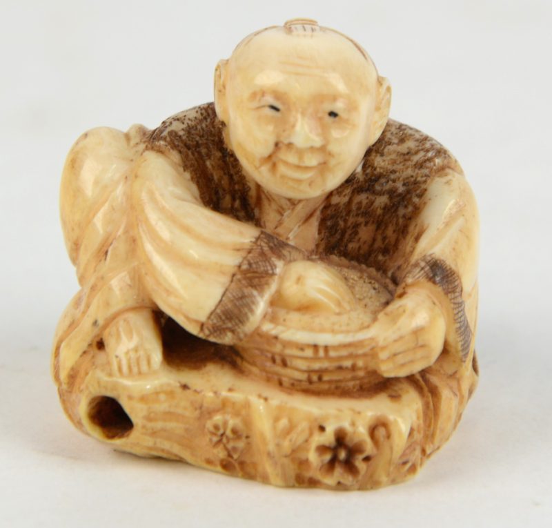 Een antieke netsuke van gesculpteerd ivoor in de vorm van een een zittende man met een mand. Onderaan gesigneerd.