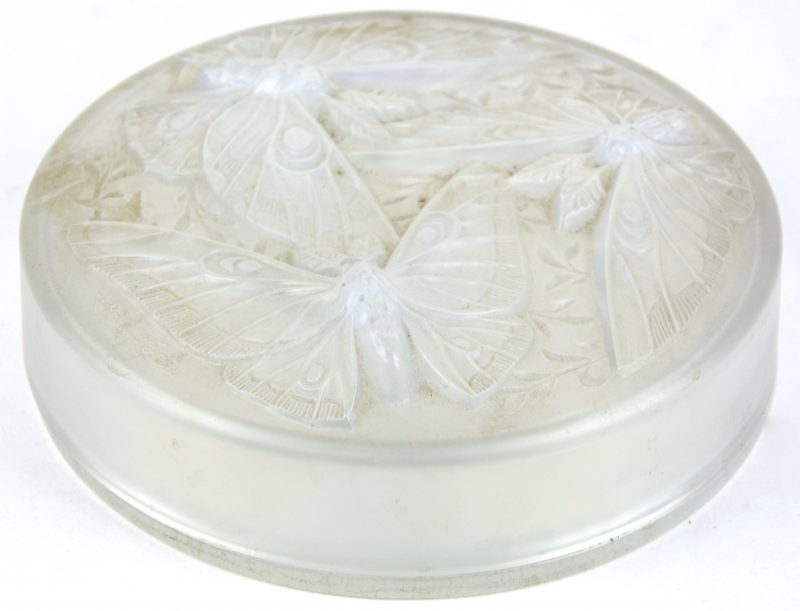 Een ronde, platte doos van iriserend glas. Het deksel versierd met drie vlinders in reliëf. Gemerkt op het deksel.