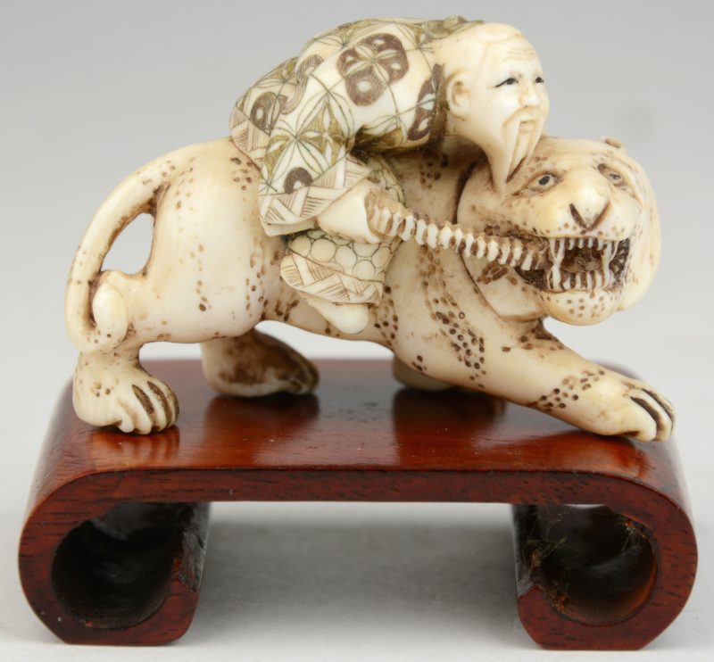 “Japans rijder op leeuw”. Een netsuke van gesculpteerd ivoor. Onderaan gesigneerd. Op houten voetstukje.