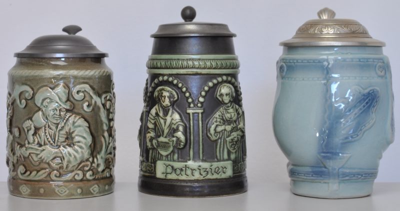 Drie bierpullen van aardewerk met tinnen deksel. Eén met een drinker in reliëf in groentinten, onderaan gemerkt Gouda (H. 15 cm). Eén met historische figuren, getiteld “Patricier”, onderaan gemerkt C.J. (H. 16,5). Eén in de vorm van een Lederhosen in lichtblauw (H. 17 cm).