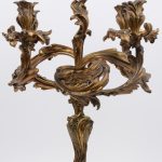 Een driedelig klokstel van verguld brons in Louis XV-stijl. De kandelaars met vijf lichtpunten in de vorm van planten en de pendule geflankeerd door twee putti. Omstreeks 1900.