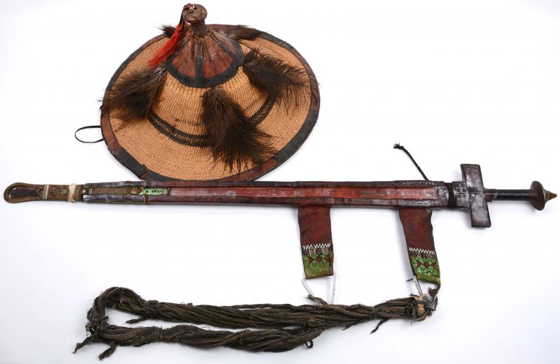 Zwaard in lederen schede en een ceremoniële hoed van raffia, afgeboord met leder en met originele pluimen. Nigeriaans werk.
