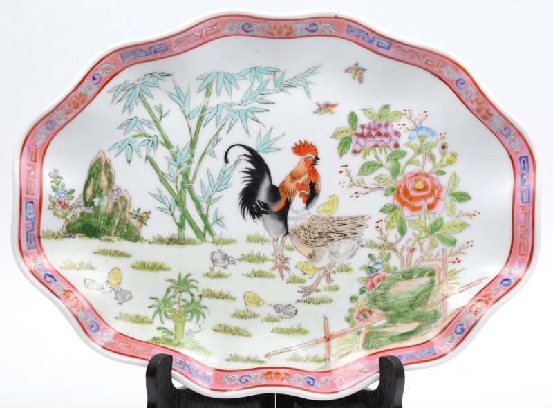 Een ovale schotel met golvende rand, versierd met een famille-rosedecor van een kip, een haan en kuikens in een tuin. Achteraan gemerkt. XXe eeuw.