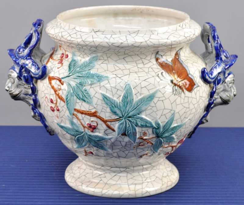 Een cachepot van meerkleurig aardewerk, versierd met een reliëf decor van  vlinders en bladeren en de handvatten in de vorm van leeuwenkoppen.