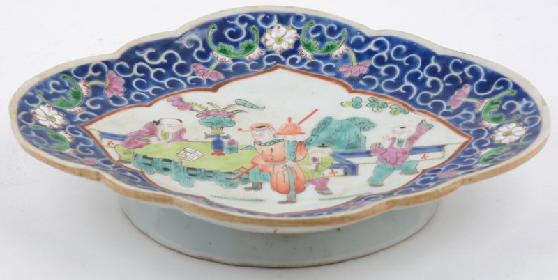 Een schaaltje op voet van meerkleurig Chinees porselein met een decor van personages en met bloemen op blauwe fond op de rand. Onderaan gemerkt.