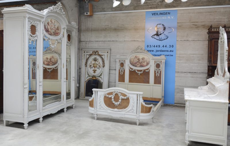 Een grijsgelakte en gesculpteerde slaapkamer in Lodewijk XVI-stijl. Bestaande uit een bed, een garderobe, een kaptafel met spiegel. Versierd met gebeeldhouwde guirlandes, met gevlochten riet en met op doek geschilderde engeltjes. Omstreeks 1900.