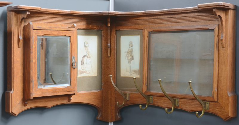 Eikenhouten art nouveau hoekkapstok met links een spiegeldeurtje, rechts vier koperen haken en een glazen opbergvak voor foto’s of brieven. Versierd met twee aquarellen gesigneerd en gedateerd 1900 en 1914.