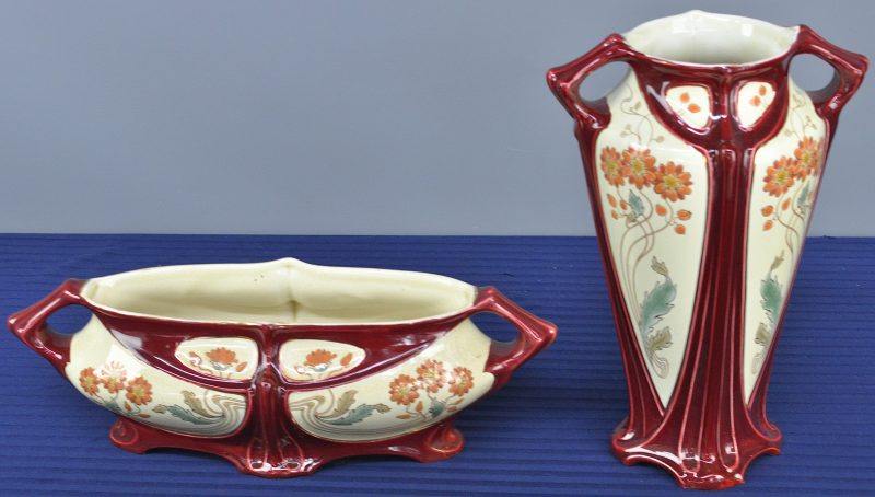 Een gedeeltelijk kaststel van meerkleurig aardewerk in art nouveaustijl, bestaande uit een coupe en een vaas, versierd met een bloemenmotief.