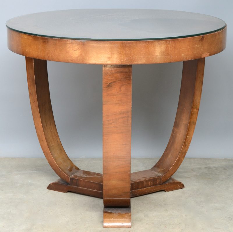 Een ronde tafel op drie gebogen poten van gefineerd hout en met een glazen blad. Tijdperk art deco.