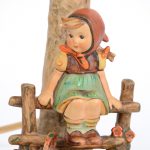Een lampvoet van meerkleurig porselein in de vorm van een meisje op een hekje bij een boomstam. Uit de reeks van M.I. Hummel. Onderaan gemerkt.