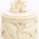 Een dekseldoos van gesculpteerd ivoor met een decor van dieren in reliëf op de zijkant en een leeuw op het deksel. Begin XXe eeuw.