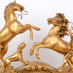 Een schouwpendule van verguld brons op onyxen voetstuk. De pendule rijkelijk versierd met classicistische voorstellingen in reliëf en bovenaan getooid met een voorstelling van een paardenmenner.