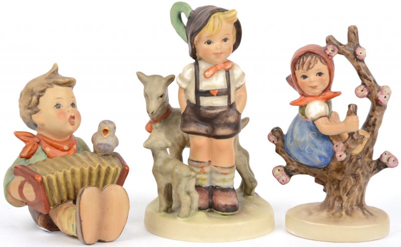 Een lot van drie beeldjes meerkleurig porselein uit de reeks van M.I. Hummel. “Jongetje met harmonica”, “Meisje op tak” en “Jongetje met twee geitjes”.