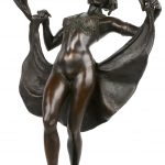 Een art deco danseres van donker gepatineerd brons met openslaand kleed op een marmeren sokkel.