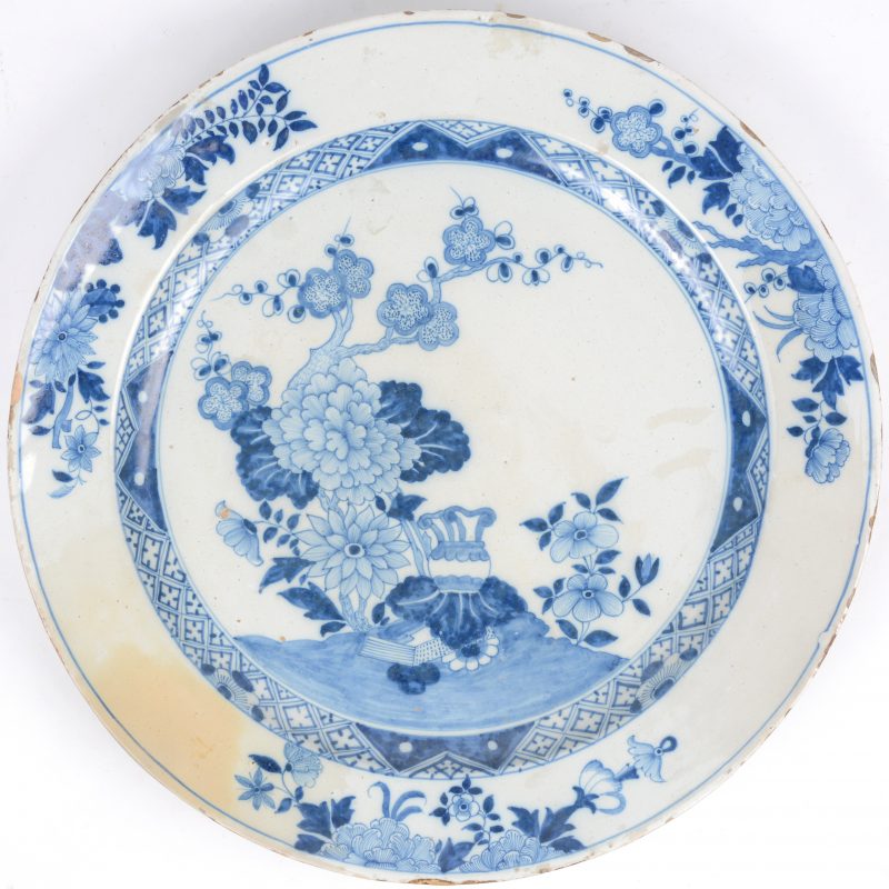 Een grote schotel van Delfts blauw aardewerk meet een Chinees decor van een bloeiende theeboom en een vaas. Gewafelde rand en  bloementuilen op de vleugel, naar Qinglong voorbeeld. Oude restauratie aan de vleugel. XVIIIde eeuw.
