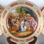 Drie schotels van polychroom porselein, met de hand beschilderd. Achteraan gemerkt en twee ervan met een opschrift: “Römischen Vasenverkaufer” en “Pyrrhäus beschutz Adromache”. Wenen, XIXde eeuw.