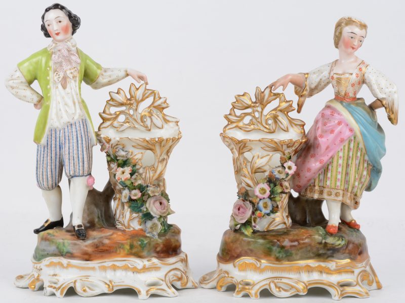 Twee groepjes van meerkleurig en verguld Frans porselein, in de vorm van een dame en een heer, telkens bij een siervaas met bloemen. Gemerkt.