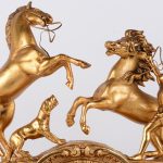 Een schouwpendule van verguld brons op onyxen voetstuk. De pendule rijkelijk versierd met classicistische voorstellingen in reliëf en bovenaan getooid met een voorstelling van een paardenmenner.