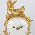 Een pendule van vuurverguld brons en wit marmer. Het uurwerk gedragen door twee engeltjes op rammen en bovenaan getooid met een drinkend naakt.