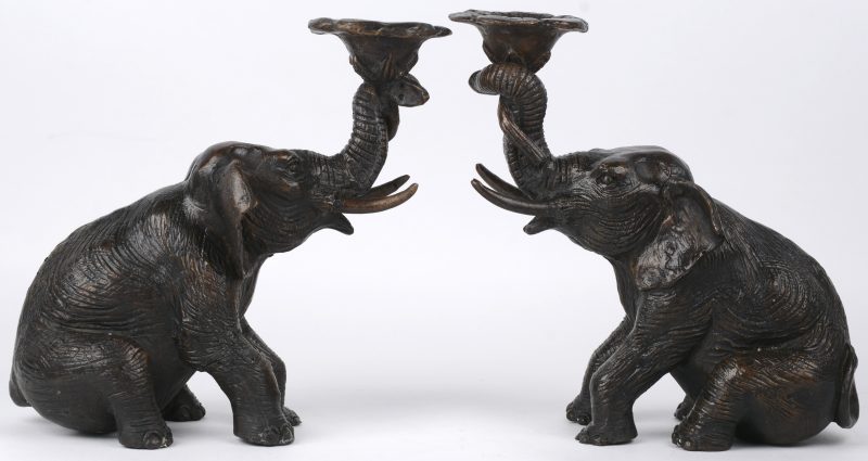 Een paar kandelaars van bruingepatineerd brons in de vorm van zittende olifanten met een bloem.