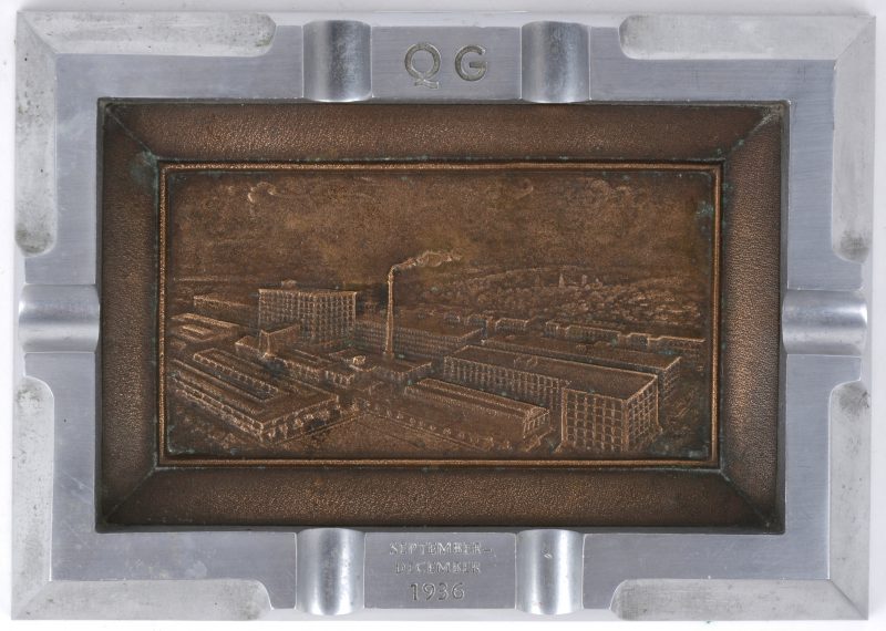 Een asbak van gepolijst metaal en koper met een reliëfafbeelding van fabrieksgebouwen. Met monogram ‘QG’ en inscriptie “September - december 1936.