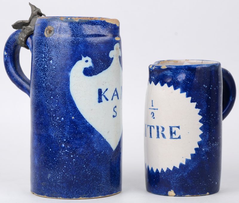 Twee oude maatbekers van blauw en wit Brussels aardewerk. Eén van een liter met opschrift ‘Kan’. Tinnen deksel manco. De andere een halve liter en XIXe eeuw.