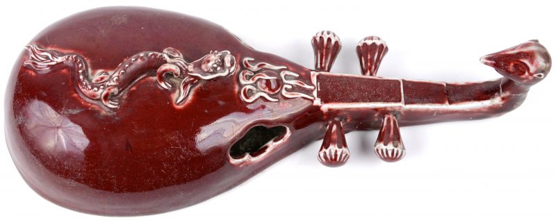 Een wandversiering van roodgeglazuurd Chinees porselein in de vorm van een snaarinstrument, versierd met een draak.