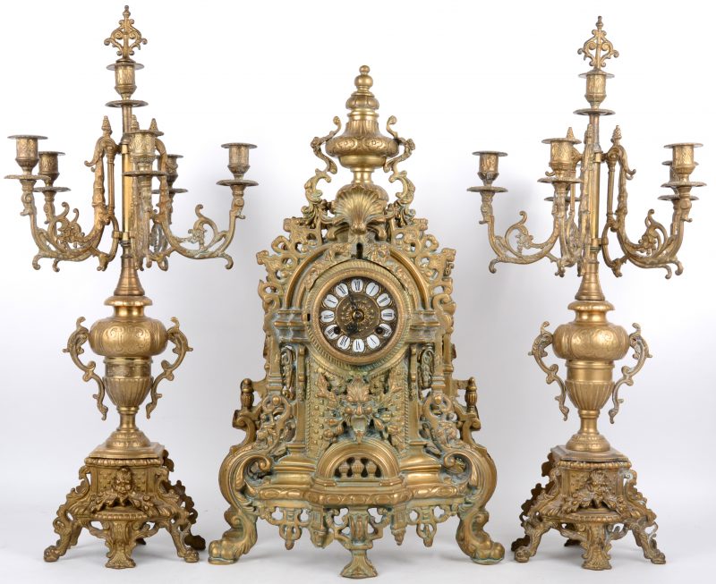 Een driedelig bronzen klokstel in barokke stijl, bestaande uit een pendule en twee kandelaars met telkens zes lichtpunten. Sleuteltje manco.