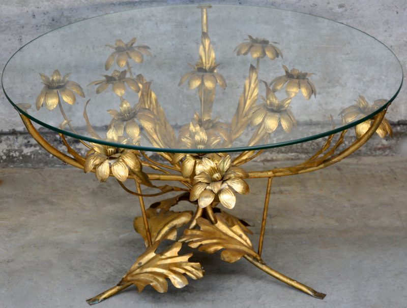 Een salontafel van goudgepatineerd smeedijzer in de vorm van bloemenranken en met een rond glazen blad.