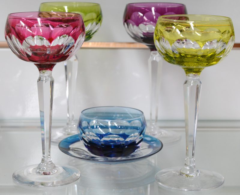 Een reeks van vier glazen op voet van geslepen kristal in verschillende kleuren. Onderaan gemerkt. We voegen er een kommetje op onderbordje in dezelfde stijl aan toe.
