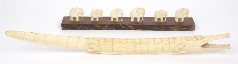 Een gesculpteerde slagtand in de vorm van een krokodil en een rijtje van zes olifntjes op houten sokkel.
