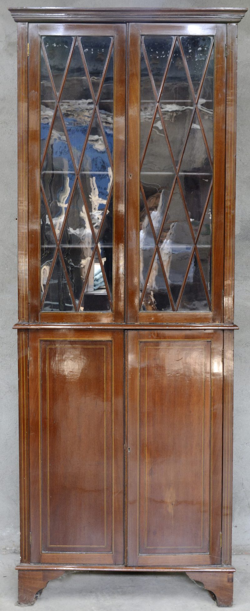 Een Engelse hoekvitrine van mahoniehoutfineer met twee paneeldeuren onderaan en versierd met een ingelegd biesje over het geheel.