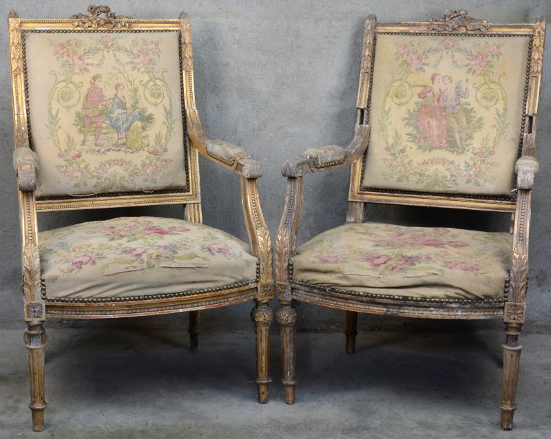Een paar armstoelen van gebeeldhouwd en verguld hout, de bekleding van naaldwerk met een decor van koppels en bloemen. Tijdperk Lodewijk XVI. Zeer sletig.