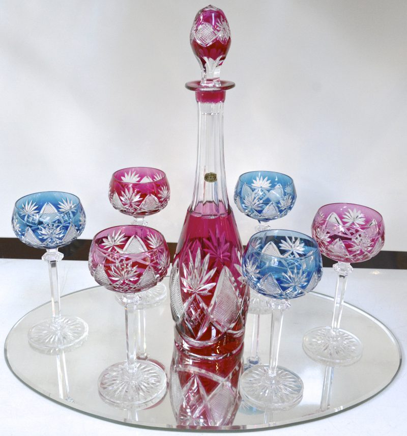 Een karaf en zes gekleurde glazen van geslepen kristal in verschillende kleuren. Op presentatiespiegel.