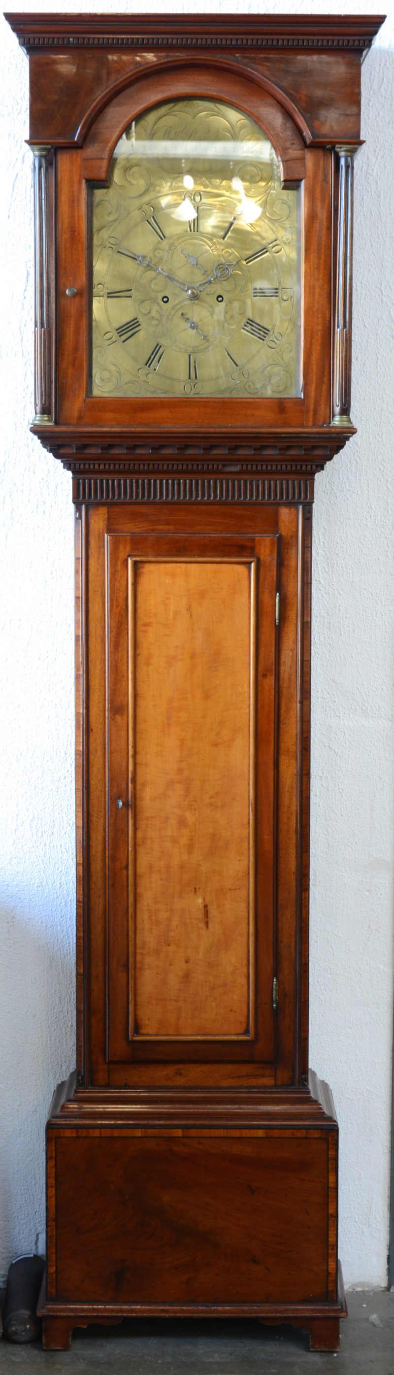 Een Engelse staande klok van mahoniehout met een gedreven messingen wijzerplaat, de kast bovenaan versierd met twee gecanneleerde zuiltjes.